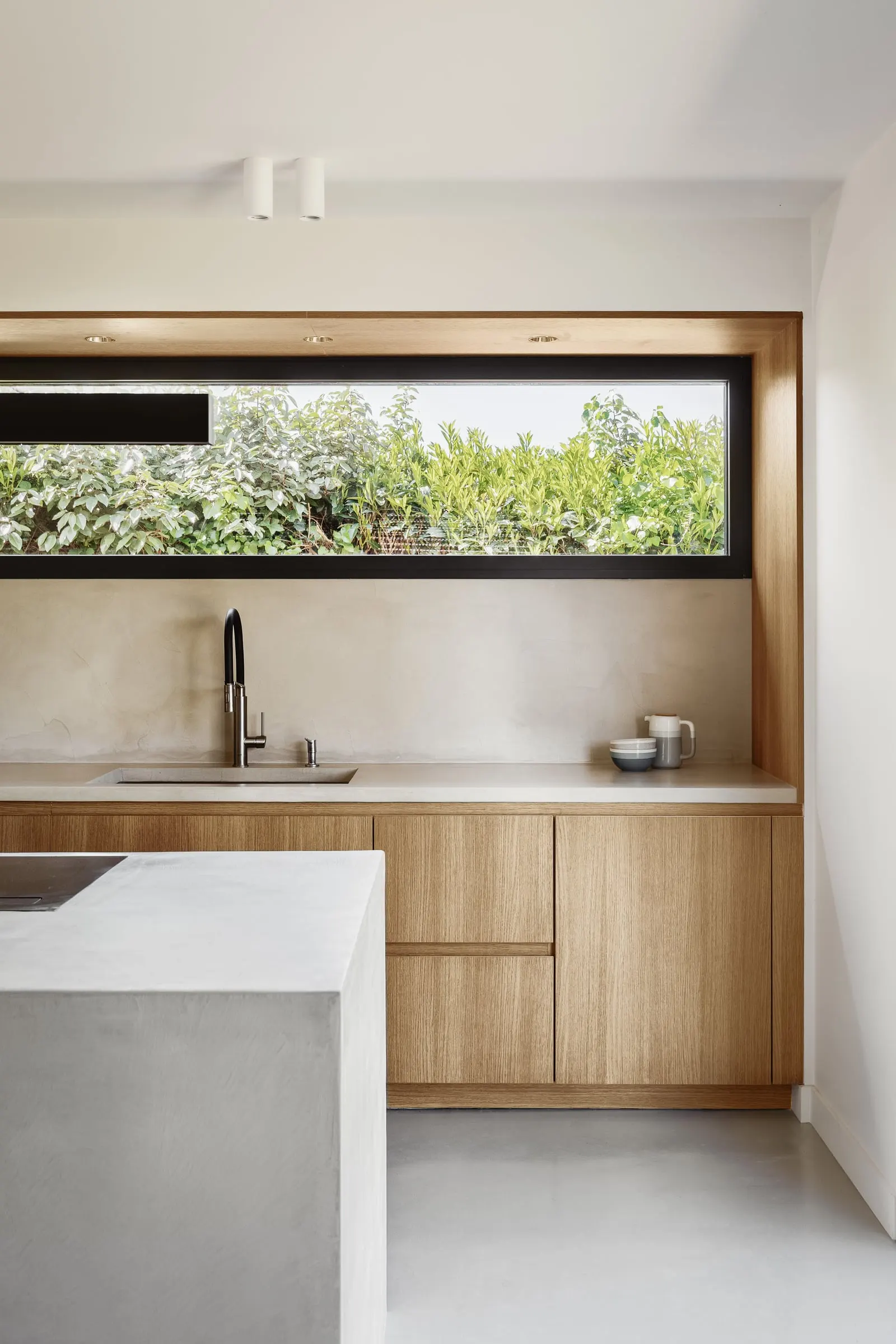 Cuisine en bois naturel et îlot minéral sous une longue fenêtre bandeau ouverte sur le jardin